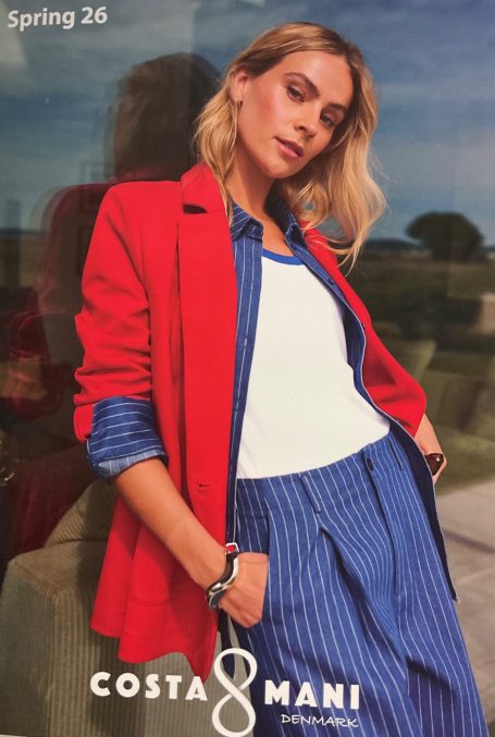 Frau in rotem Blazer und gestreifter Bluse posiert entspannt vor blauem Hintergrund.